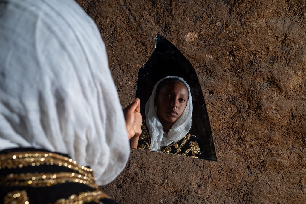 Mart is wearing traditional wedding clothing in a provocative protest against child marriage. Her attire is a white dress with red trim and a black and gold cape. She is looking at herself in a mirror shard.